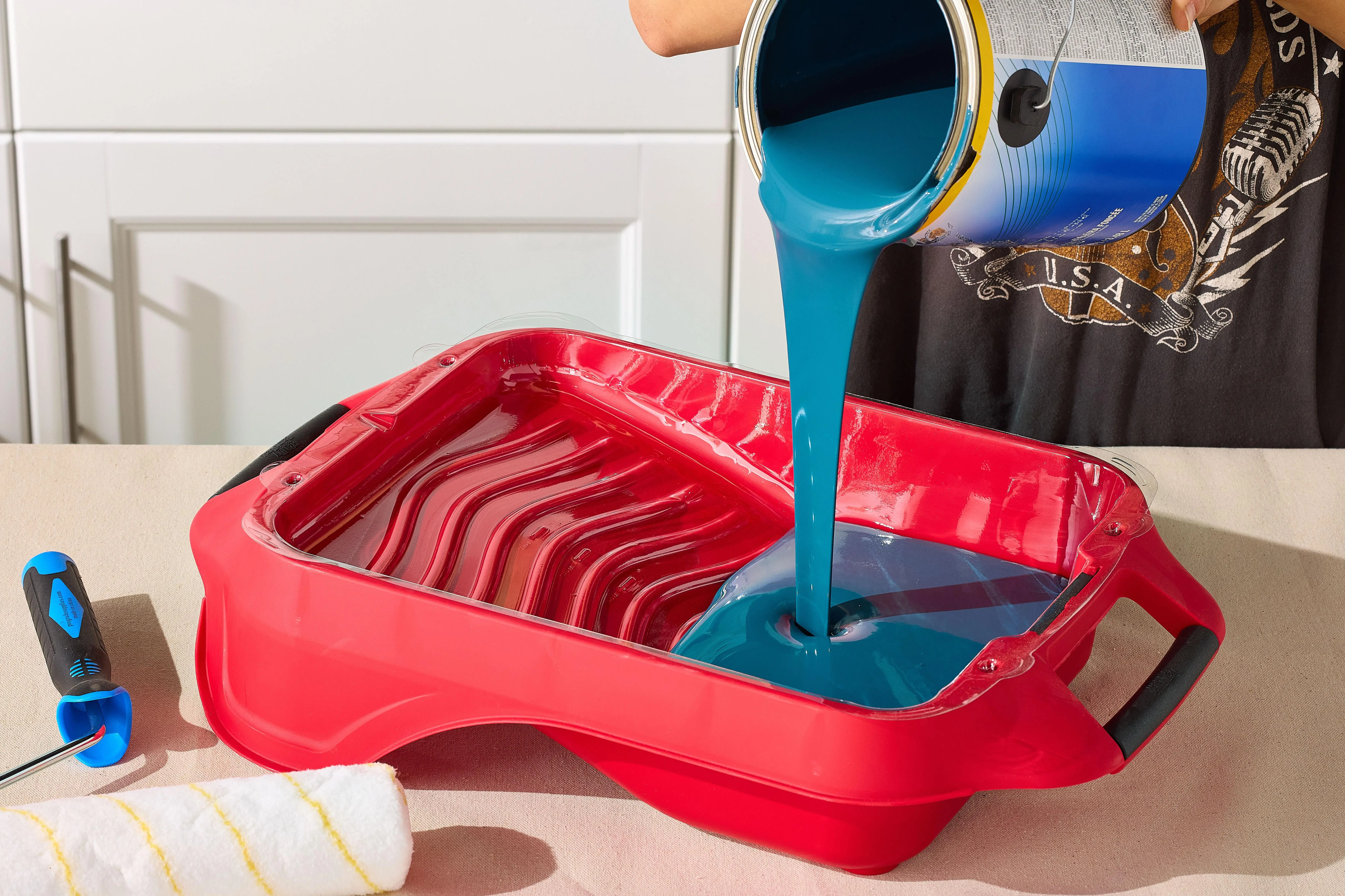 Blue latex paint being poured from a can into a red paint tray, preparing for an interior house painting project.